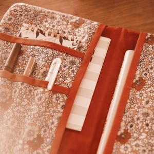 May include: A floral patterned notebook cover with a red border. It contains a spiral-bound notebook, pens, and a notepad. The floral pattern has white and orange flowers on a brown background.