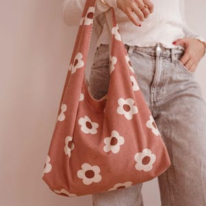 May include: A pink corduroy shoulder bag with a white daisy pattern. The bag has a wide strap and a simple, casual design. The bag is a soft, muted pink colour.
