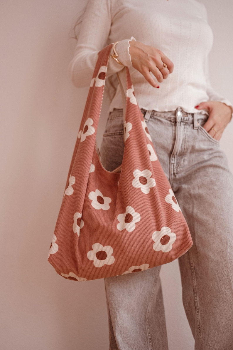 May include: A pink corduroy shoulder bag with a white daisy pattern. The bag has a wide strap and a simple, casual design. The bag is a soft, muted pink colour.