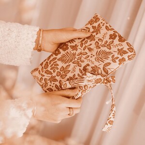 May include: A beige clutch with a brown floral pattern, held by hands. The clutch features a bow closure and a matching strap. The floral design includes leaves and flowers. The background is a soft, blurred neutral colour.