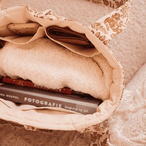May include: Open tote bag with a floral pattern in shades of beige and brown. The bag is filled with a white towel, a brown wallet, a wooden item, and a book with the title "FOTOGRAFIE Inspired by Life".