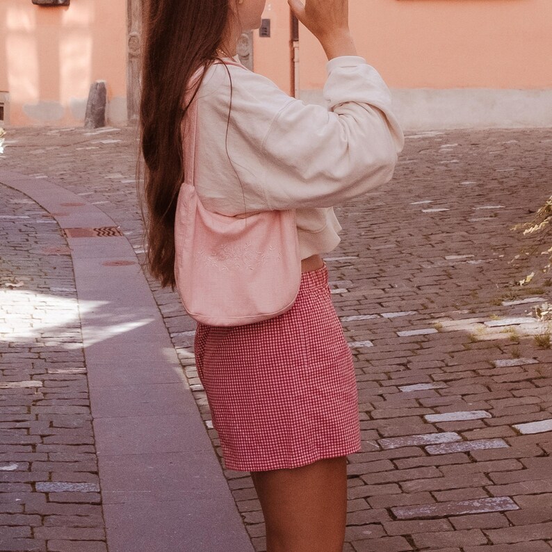 May include: A light pink hobo-style shoulder bag with a single strap, worn over a cream-colored long-sleeve top and red and white gingham patterned shorts. The bag features a soft, unstructured silhouette and a smooth, solid-colored exterior finish.