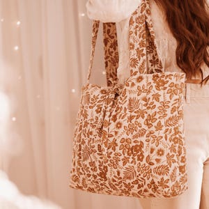 May include: A beige tote bag with a brown floral pattern. The bag has long straps and is held over the shoulder. The floral design includes leaves and flowers.