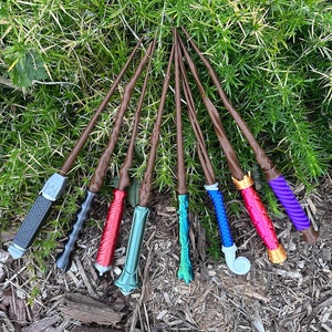 以下が含まれることがあります： 茶色の木製の軸とカラフルな塗装された先端を持つ、8本の異なる魔法の杖。杖は、緑の葉と茶色のマルチのベッドの上に扇形に配置されています。