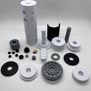 Puede incluir: Surtido de piezas impresas en 3D en blanco, negro y gris. Las piezas incluyen un cilindro alto con agujeros, una pieza curva, un cilindro peque&ntilde;o, un disco redondo con agujeros y varias piezas m&aacute;s peque&ntilde;as.