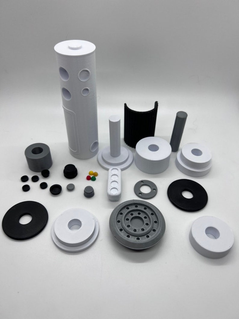 Puede incluir: Surtido de piezas impresas en 3D en blanco, negro y gris. Las piezas incluyen un cilindro alto con agujeros, una pieza curva, un cilindro peque&ntilde;o, un disco redondo con agujeros y varias piezas m&aacute;s peque&ntilde;as.