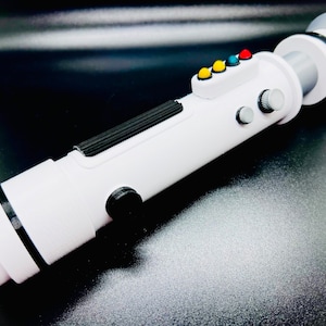 Puede incluir: Juguete de sable de luz blanco con detalles negros y botones de colores. El sable de luz tiene una superficie texturizada y un anillo negro alrededor del extremo.