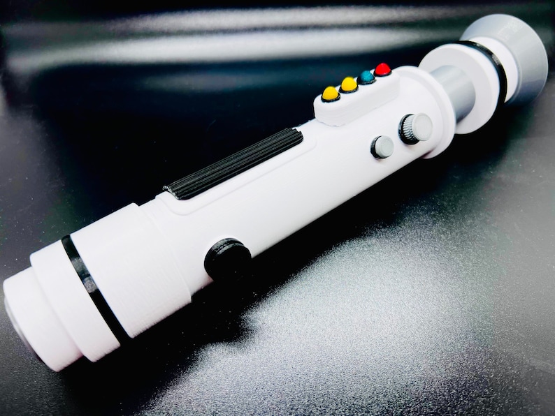 Puede incluir: Juguete de sable de luz blanco con detalles negros y botones de colores. El sable de luz tiene una superficie texturizada y un anillo negro alrededor del extremo.