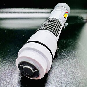 Puede incluir: Accesorio de sable de luz blanco con detalles negros y grises. El sable de luz tiene un bot&oacute;n negro en el extremo y un peque&ntilde;o bot&oacute;n de colores en el lateral.