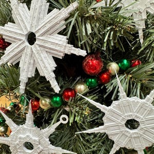 May include: White snowflake-shaped Christmas ornaments with a detailed design, hanging on a green Christmas tree with red and green ornaments and a silver and gold beaded garland.