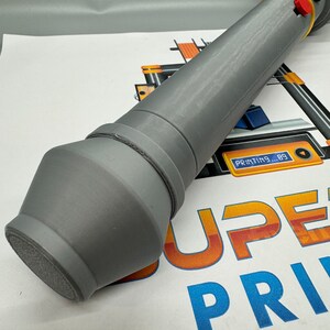 May include: A grey 3D printed object with a rounded end and a yellow and red button on the top. The object is lying on a white surface with the text "SUPER PRINT" in orange and blue.