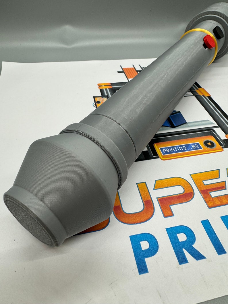 May include: A grey 3D printed object with a rounded end and a yellow and red button on the top. The object is lying on a white surface with the text "SUPER PRINT" in orange and blue.
