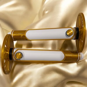 K&ouml;nnte beinhalten: Zwei dekorative, gold- und wei&szlig;farbene Objekte mit einem rad&auml;hnlichen Design an jedem Ende. Die Objekte haben einen wei&szlig;en Mittelteil mit goldenen Akzenten und ein kleines, kreisf&ouml;rmiges Golddetail. Der Hintergrund ist ein goldfarbener Satin-Stoff.