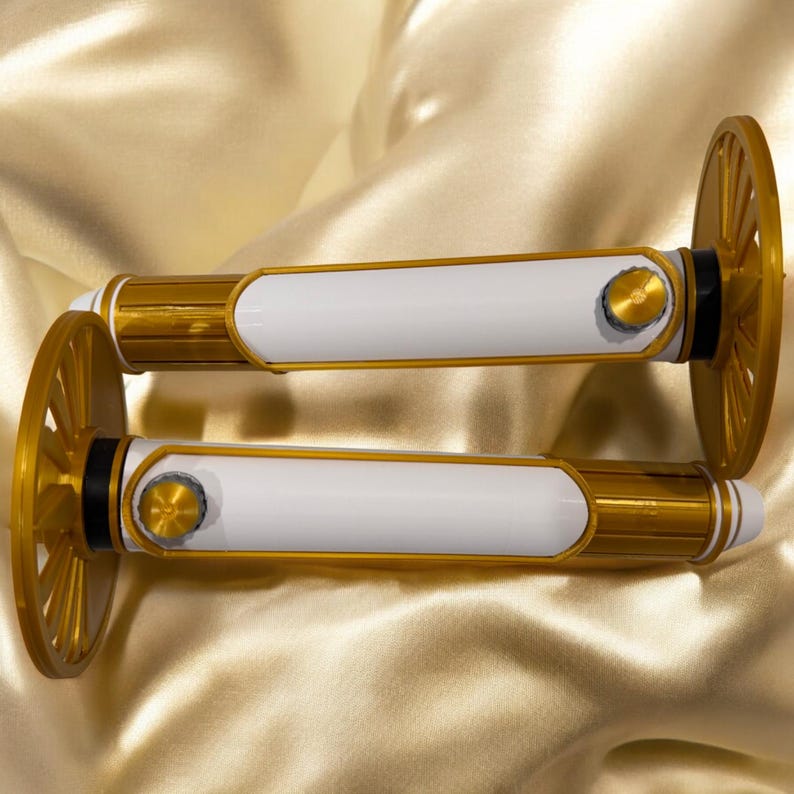 K&ouml;nnte beinhalten: Zwei dekorative, gold- und wei&szlig;farbene Objekte mit einem rad&auml;hnlichen Design an jedem Ende. Die Objekte haben einen wei&szlig;en Mittelteil mit goldenen Akzenten und ein kleines, kreisf&ouml;rmiges Golddetail. Der Hintergrund ist ein goldfarbener Satin-Stoff.