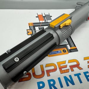 May include: A gray 3D printed lightsaber with a yellow button and black accents. The lightsaber is resting on a white surface with the text "SUPER 3D PRINTER" printed on it.