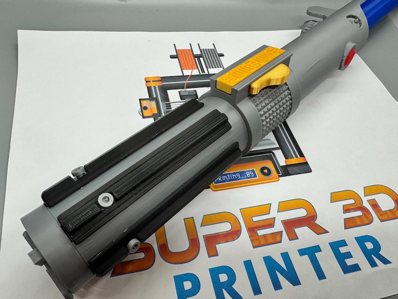 May include: A gray 3D printed lightsaber with a yellow button and black accents. The lightsaber is resting on a white surface with the text "SUPER 3D PRINTER" printed on it.