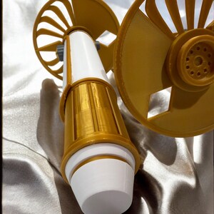 K&ouml;nnte beinhalten: Ein 3D-gedrucktes Ger&auml;t in Wei&szlig; und Gold, mit zwei f&auml;cherf&ouml;rmigen Elementen und einem zylindrischen K&ouml;rper. Das Design kombiniert glatte und strukturierte Oberfl&auml;chen. Das Ger&auml;t hat eine futuristische &Auml;sthetik.
