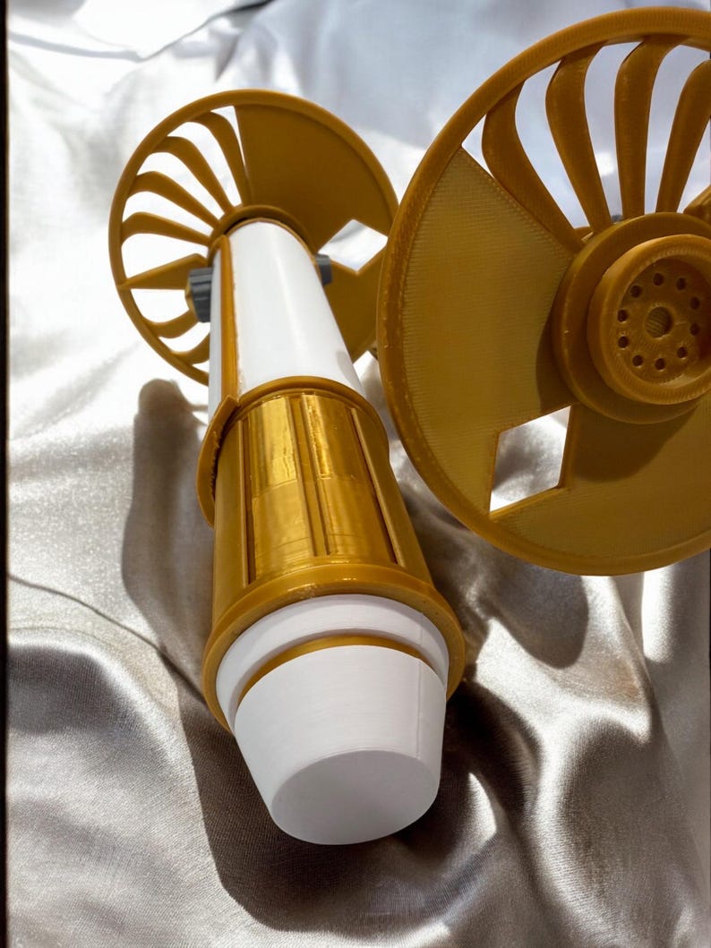 K&ouml;nnte beinhalten: Ein 3D-gedrucktes Ger&auml;t in Wei&szlig; und Gold, mit zwei f&auml;cherf&ouml;rmigen Elementen und einem zylindrischen K&ouml;rper. Das Design kombiniert glatte und strukturierte Oberfl&auml;chen. Das Ger&auml;t hat eine futuristische &Auml;sthetik.