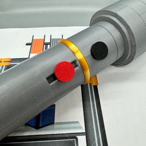May include: A grey 3D printed object with a gold band and two red and black circular buttons. The object is being printed on a 3D printer with a yellow and black display that reads "PRINTING... 89".