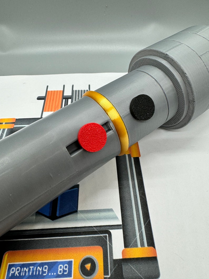 May include: A grey 3D printed object with a gold band and two red and black circular buttons. The object is being printed on a 3D printer with a yellow and black display that reads "PRINTING... 89".