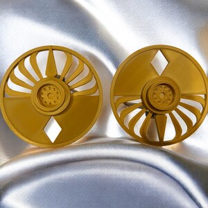 K&ouml;nnte beinhalten: Zwei goldfarbene, kreisf&ouml;rmige Ohrringe mit geometrischem Design. Jeder Ohrring hat einen detaillierten Mittelkreis, der von gebogenen, f&auml;cherartigen Elementen und einem rautenf&ouml;rmigen Ausschnitt umgeben ist. Die Ohrringe werden auf einem gl&auml;nzenden, silbernen Stoff pr&auml;sentiert.