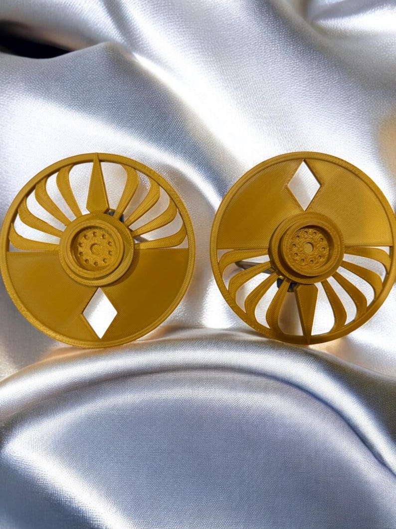 K&ouml;nnte beinhalten: Zwei goldfarbene, kreisf&ouml;rmige Ohrringe mit geometrischem Design. Jeder Ohrring hat einen detaillierten Mittelkreis, der von gebogenen, f&auml;cherartigen Elementen und einem rautenf&ouml;rmigen Ausschnitt umgeben ist. Die Ohrringe werden auf einem gl&auml;nzenden, silbernen Stoff pr&auml;sentiert.