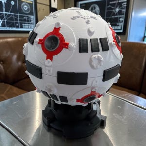 May include: A white, spherical, 3D-printed model with black and red accents. The model features rectangular and circular details, and sits on a black base. The design is reminiscent of a futuristic or sci-fi aesthetic.