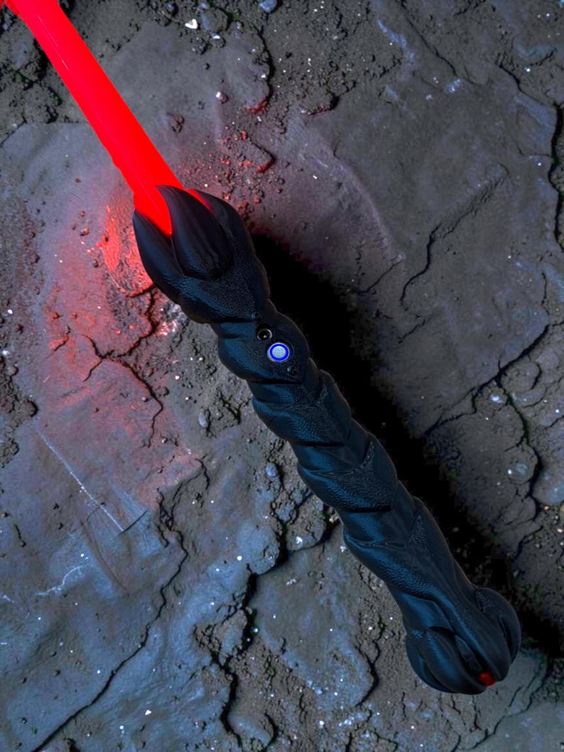 May include: A black lightsaber with a textured handle and a glowing red blade. The hilt features a blue button and a claw-like design at the blade's base. The lightsaber is set against a dark, textured background.
