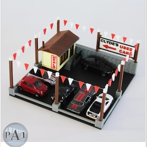 May include: A miniature model of a used car dealership with a small building, a sign that reads "Clyde's Used Cars", and several toy cars in the lot. The dealership is decorated with red and white bunting.