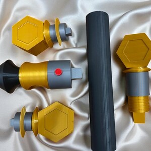 May include: A collection of 3D-printed items in gold, gray, and black. The items include a long cylinder and several pieces with hexagonal and cylindrical shapes. One piece has a red button.