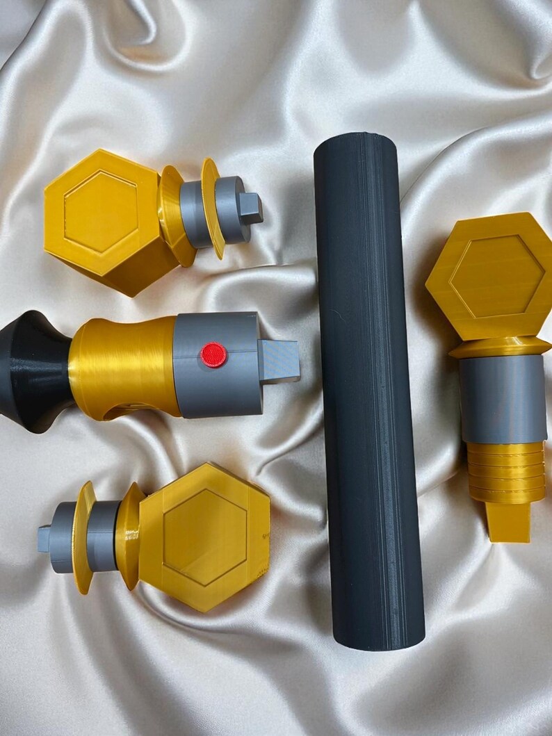 May include: A collection of 3D-printed items in gold, gray, and black. The items include a long cylinder and several pieces with hexagonal and cylindrical shapes. One piece has a red button.