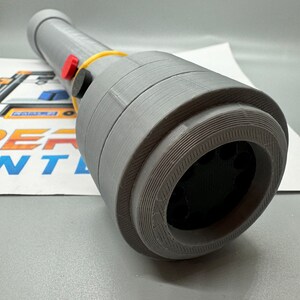 May include: A grey 3D printed object with a black circular opening and a yellow rubber band. There are two red buttons and a black button on the side of the object.