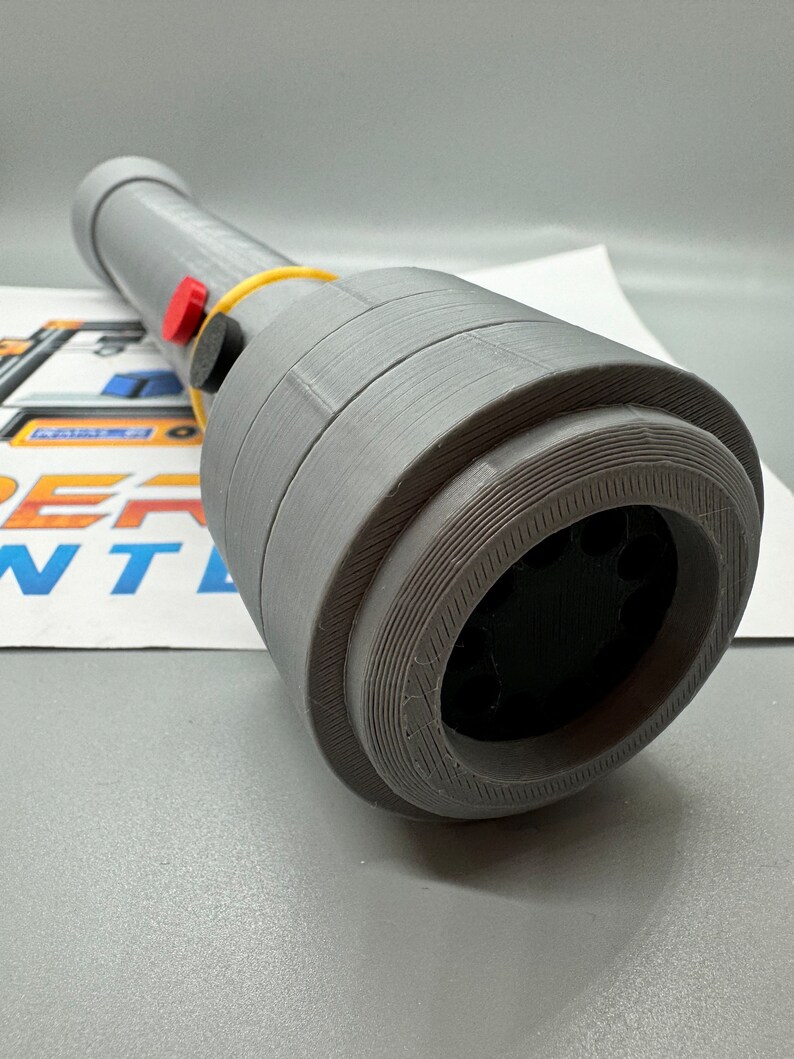 May include: A grey 3D printed object with a black circular opening and a yellow rubber band. There are two red buttons and a black button on the side of the object.