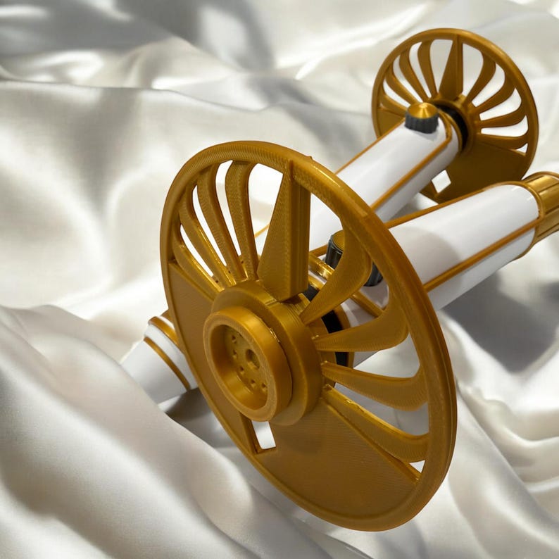 K&ouml;nnte beinhalten: Ein dekoratives Kanonenmodell mit goldfarbenen R&auml;dern und Akzenten sowie wei&szlig;en L&auml;ufen. Die R&auml;der haben ein Speichendesign. Die Kanone ist vor einem wei&szlig;en, seidigen Hintergrund platziert.