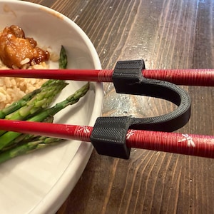 以下が含まれることがあります： 食べ物の皿の上に置かれた、黒い箸置きが付いた赤い箸。箸には赤と白の花柄のデザインがあります。お皿には玄米、アスパラガス、鶏肉が入っています。