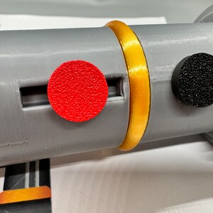 May include: A close-up of a grey cylindrical object with a gold band around it. There is a red circular button on the left and a black circular button on the right.