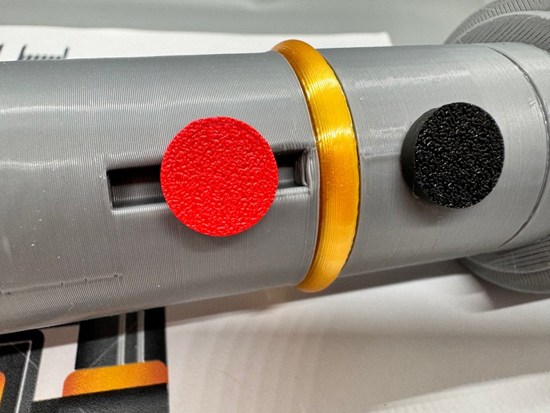 May include: A close-up of a grey cylindrical object with a gold band around it. There is a red circular button on the left and a black circular button on the right.