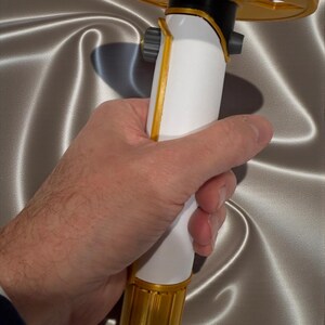 K&ouml;nnte beinhalten: Ein wei&szlig;-goldenes Lichtschwert-Requisit mit einem kreisf&ouml;rmigen goldenen Knauf. Das Lichtschwert hat einen wei&szlig;en Griff mit goldenen Akzenten und einen schwarzen Abschnitt in der N&auml;he des Knaufs. Die Requisite wird in einer Hand vor einem silbernen Satinhintergrund gehalten.