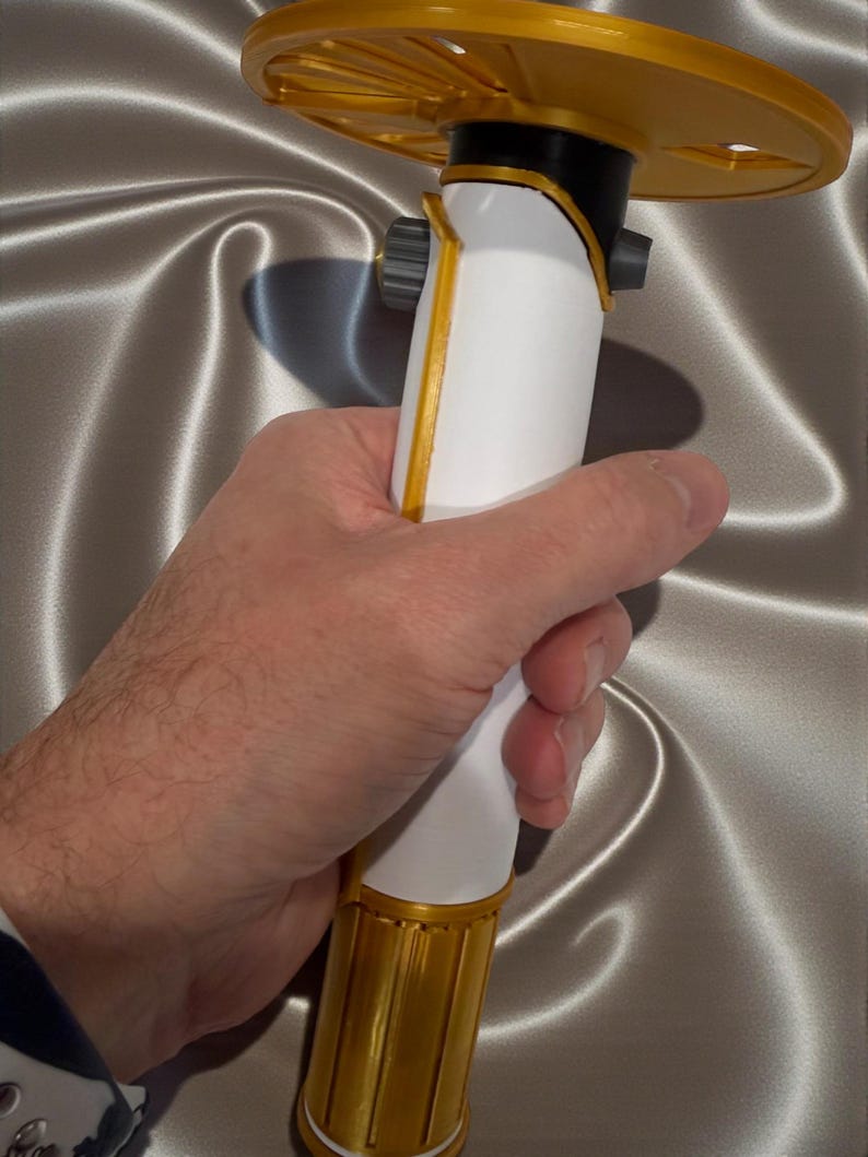 K&ouml;nnte beinhalten: Ein wei&szlig;-goldenes Lichtschwert-Requisit mit einem kreisf&ouml;rmigen goldenen Knauf. Das Lichtschwert hat einen wei&szlig;en Griff mit goldenen Akzenten und einen schwarzen Abschnitt in der N&auml;he des Knaufs. Die Requisite wird in einer Hand vor einem silbernen Satinhintergrund gehalten.