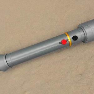May include: A grey plastic toy lightsaber with a gold and red button. The lightsaber is lying on a sandy surface.