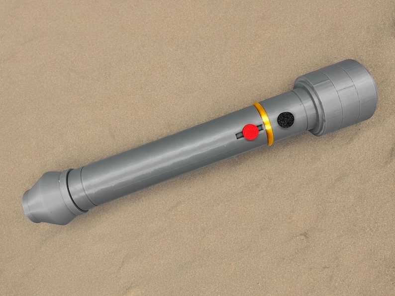 May include: A grey plastic toy lightsaber with a gold and red button. The lightsaber is lying on a sandy surface.