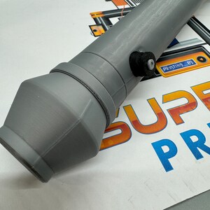 May include: A grey 3D printed nozzle with a black knob on the side. The nozzle is designed for a 3D printer and has a round opening at the end.