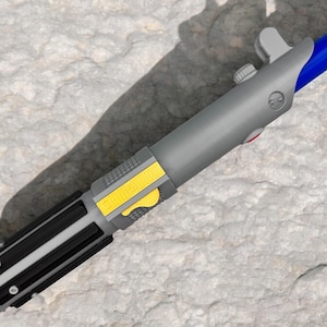 Puede incluir: Un juguete de sable de luz gris con un mango negro y un botón amarillo. El sable de luz tiene una hoja azul que no es visible en la imagen.