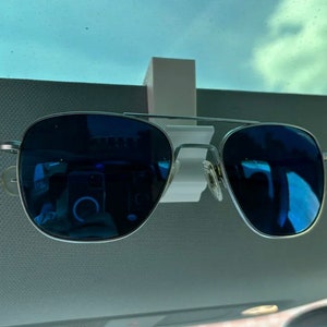 May include: A pair of blue aviator sunglasses are held in place by a white plastic clip attached to a car's sun visor.