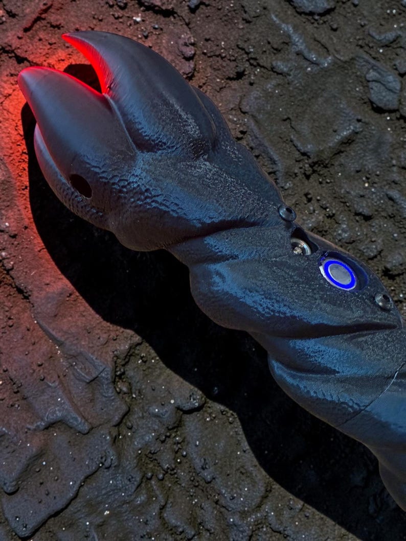 May include: A dark blue, claw-shaped object with a textured surface and a red glowing tip. The object has a series of small, circular details and a blue illuminated circle. The object is set against a dark, textured background.