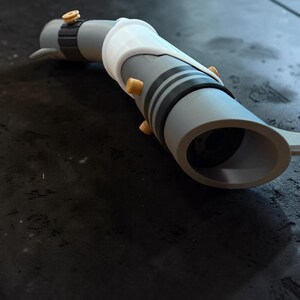 K&ouml;nnte beinhalten: Eine graue und wei&szlig;e Lichtschwert-Requisite mit schwarzen Streifen und goldfarbenen Akzenten. Die Requisite hat ein zylindrisches Design mit einem wei&szlig;en Griff und einer kreisf&ouml;rmigen &Ouml;ffnung am Ende. Das Lichtschwert liegt auf einer dunklen Oberfl&auml;che.