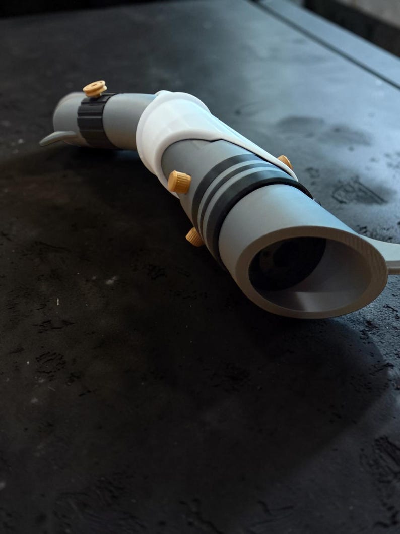K&ouml;nnte beinhalten: Eine graue und wei&szlig;e Lichtschwert-Requisite mit schwarzen Streifen und goldfarbenen Akzenten. Die Requisite hat ein zylindrisches Design mit einem wei&szlig;en Griff und einer kreisf&ouml;rmigen &Ouml;ffnung am Ende. Das Lichtschwert liegt auf einer dunklen Oberfl&auml;che.