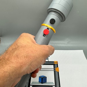 May include: A grey 3D printed object with a black and red button. The object is being held in a person's hand. The object is being printed on a 3D printer. The printer's display shows "PRINTING... 89".