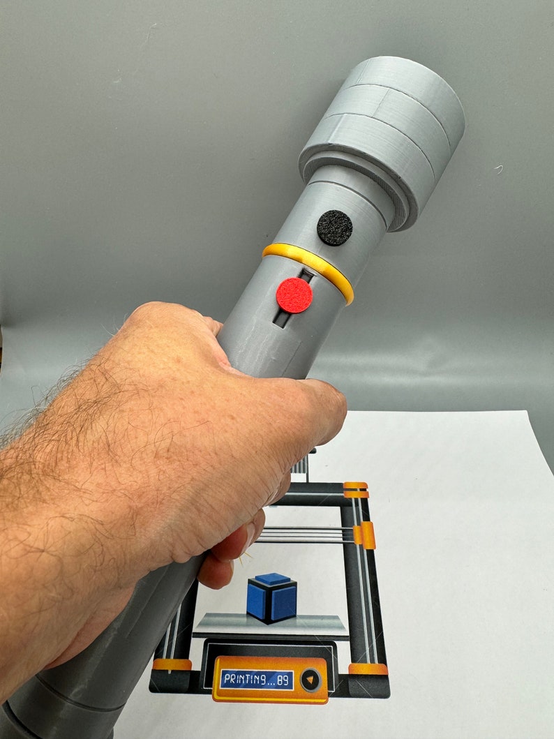 May include: A grey 3D printed object with a black and red button. The object is being held in a person's hand. The object is being printed on a 3D printer. The printer's display shows "PRINTING... 89".