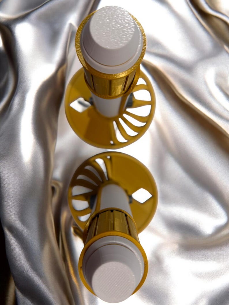 K&ouml;nnte beinhalten: Zwei dekorative Flaschenverschl&uuml;sse. Jeder Verschluss hat eine wei&szlig;e Oberseite mit strukturierter Oberfl&auml;che, ein goldfarbenes Band und eine gelbe Basis mit ausgeschnittenen Designs. Die Verschl&uuml;sse sind vor einem silbernen Satinhintergrund platziert.